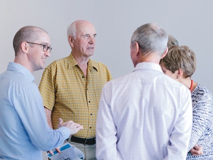 A group of five adults is having a conversation in a bright room. One man on the left is holding a notebook, while an older man in a yellow checkered shirt stands in the center. The other three are facing away, engaged in the discussion.