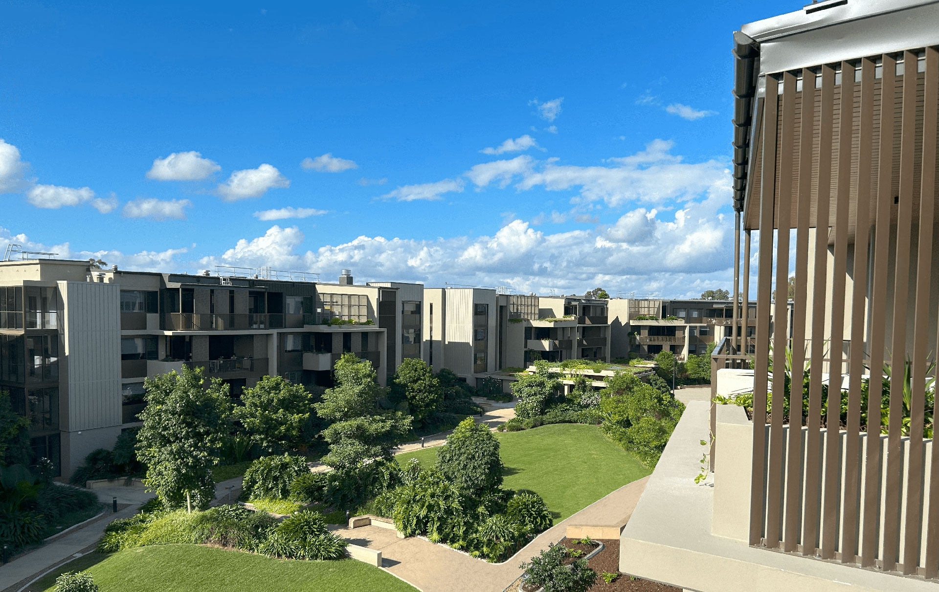 A modern apartment complex with multiple buildings surrounded by lush greenery and well-maintained lawns under a clear blue sky with scattered clouds. A balcony with vertical slats is visible on the right.