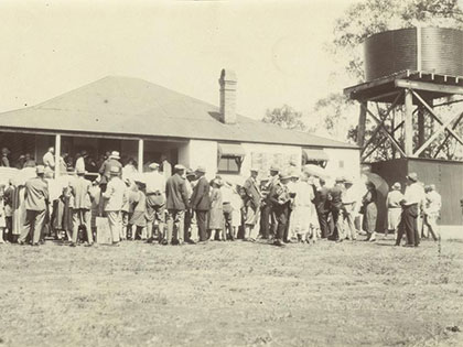 A historical black and white photo shows a crowd of people gathered outside a building with a chimney. A large water tank on a stand is visible to the right. The scene suggests a community event or gathering.