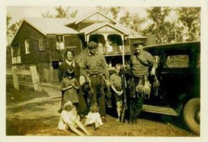 Vintage photo of a family in front of a wooden house and an old car. Two men and a woman stand with children and dolls, depicting a rustic scene from the past. Trees and a wooden fence surround the home.