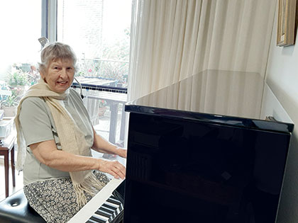An elderly woman with gray hair plays the piano, wearing a light scarf and short-sleeved top. She is seated on a bench, and a window with potted plants is visible in the background.