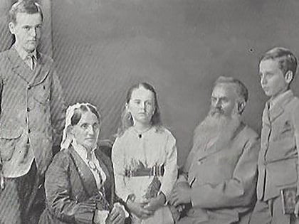 A vintage black and white photo of a family group. An older couple seated, with a bearded man and a woman wearing a bonnet. Three children stand around them, two boys in suits and a girl in a dress, all looking towards the camera.