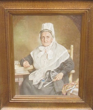 A portrait of an elderly woman seated on a chair, dressed in traditional attire with a white headpiece and shawl. She holds a small book and pen, with a subtle smile, set against a neutral background in a wooden frame.