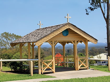 A small wooden gazebo with a shingled roof, featuring two crosses on top. The structure is surrounded by a grassy area and trees, with a clear sky and mountains visible in the background.