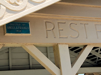 Close-up of a wooden beam structure with carved text partially visible, reading "REST F." A small blue plaque reads "Erected by Angela Francis 1902." The structure has decorative elements and is painted in a light color.