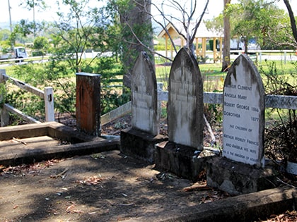 An old cemetery with three weathered tombstones in a rural setting, surrounded by greenery. A white wooden fence separates the graves from the background, which features a small house and a road. Sunlight filters through the trees.