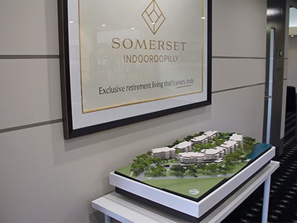 A scale model of a modern retirement living complex is displayed on a table. Above it, a framed sign reads "Somerset Indooroopilly: Exclusive retirement living that's yours, truly." The interior space is well-lit and contemporary.
