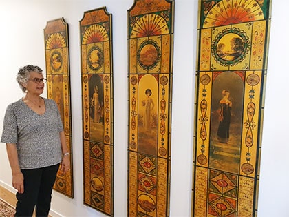 A person stands in a gallery observing four tall, narrow paintings with ornate designs and classical figures on a wall. Each artwork features warm tones and intricate patterns with a central figure. The atmosphere is calm and contemplative.