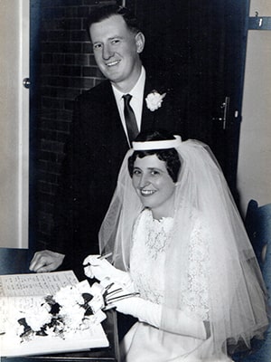 A black and white photo of a bride and groom on their wedding day. The bride is seated, wearing a lace wedding gown and veil, smiling and holding a pen over a document. The groom stands beside her in a dark suit with a flower boutonniere, smiling.