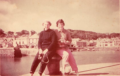 Two people sit on a dock by the water, with a scenic view of buildings and trees in the background. The image has a vintage, sepia tone, giving it an old-fashioned feel.