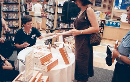 A woman in a brown dress is handing a book to a seated person at a book signing event in a bookstore. A table is covered with stacks of books, and other people browse in the background.