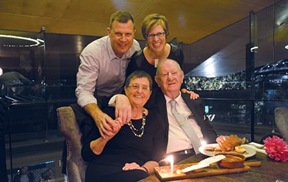 Four people are gathered around a table in a restaurant setting. Two are seated, while two stand behind them, all smiling. The table has a small cake with lit candles. The background includes modern decor with a glass balcony.