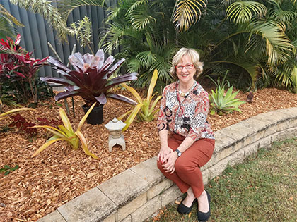 A woman with short hair and glasses sits on a low stone wall in a garden. She is wearing a patterned blouse and orange pants. The garden features various plants, including a large bromeliad and ferns, with a small lantern decoration nearby.
