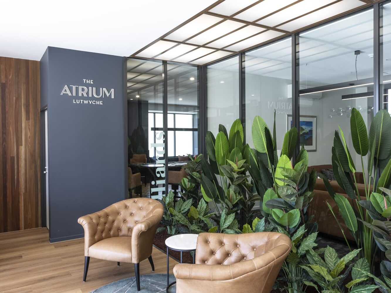 Modern waiting area with two brown leather chairs, a small round table on a geometric-patterned rug, large potted plants, and a wooden floor. A wall sign reads "The Atrium Lutwyche." Natural light filters through a large window.