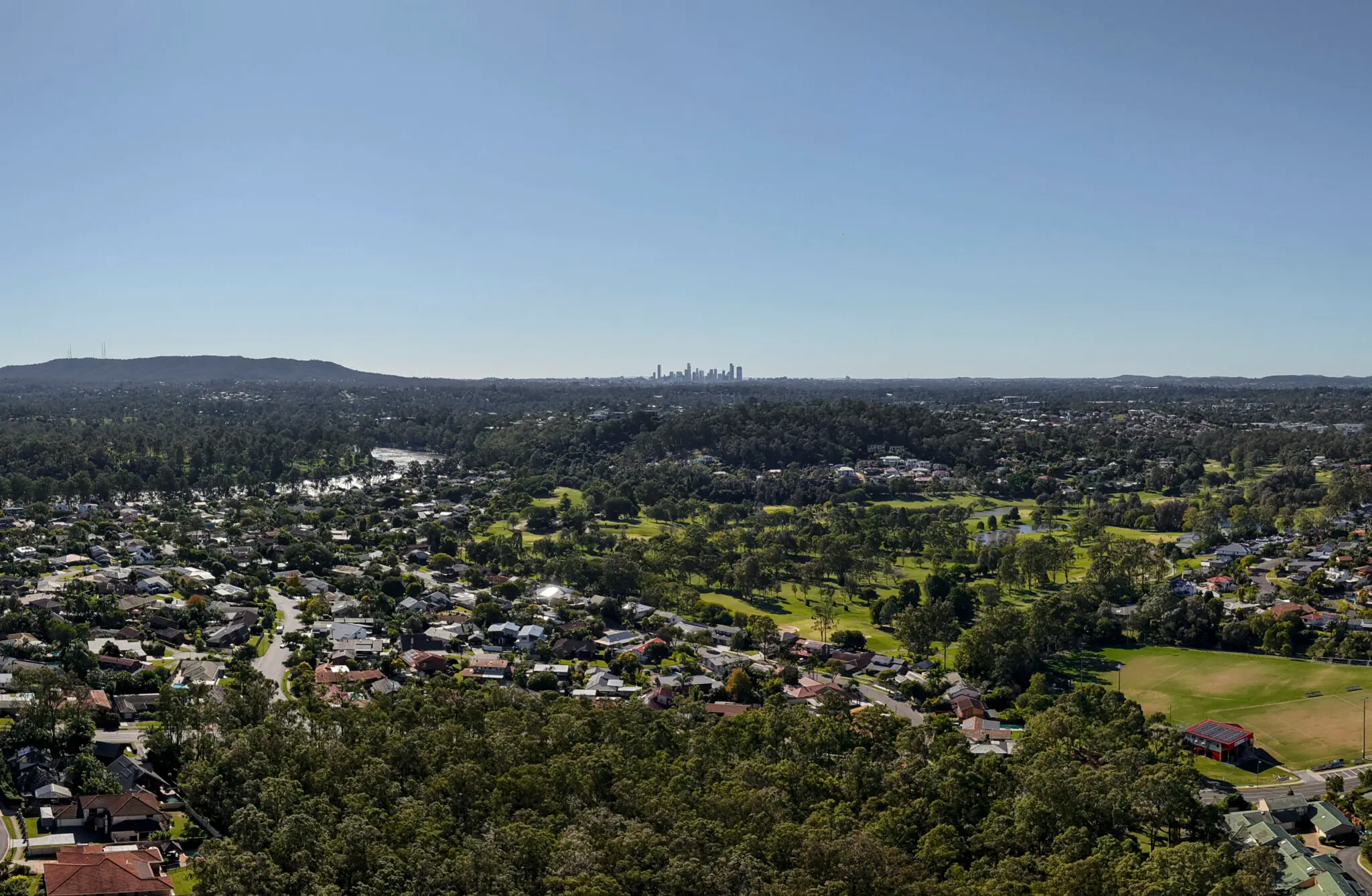 A panoramic view of a suburban area with many trees, houses, and green spaces under a clear blue sky. In the distance, The Evelyn Mt Ommaney rises near the city's skyline on the horizon.
