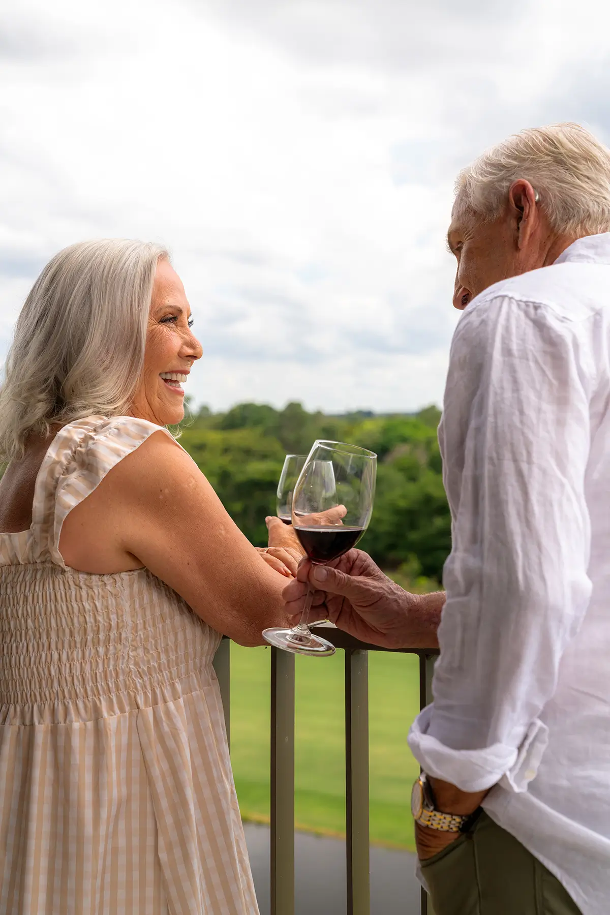 An older couple stands on a balcony at The Evelyn Mt Ommaney, smiling and holding glasses of red wine, with a green landscape and cloudy sky in the background.
