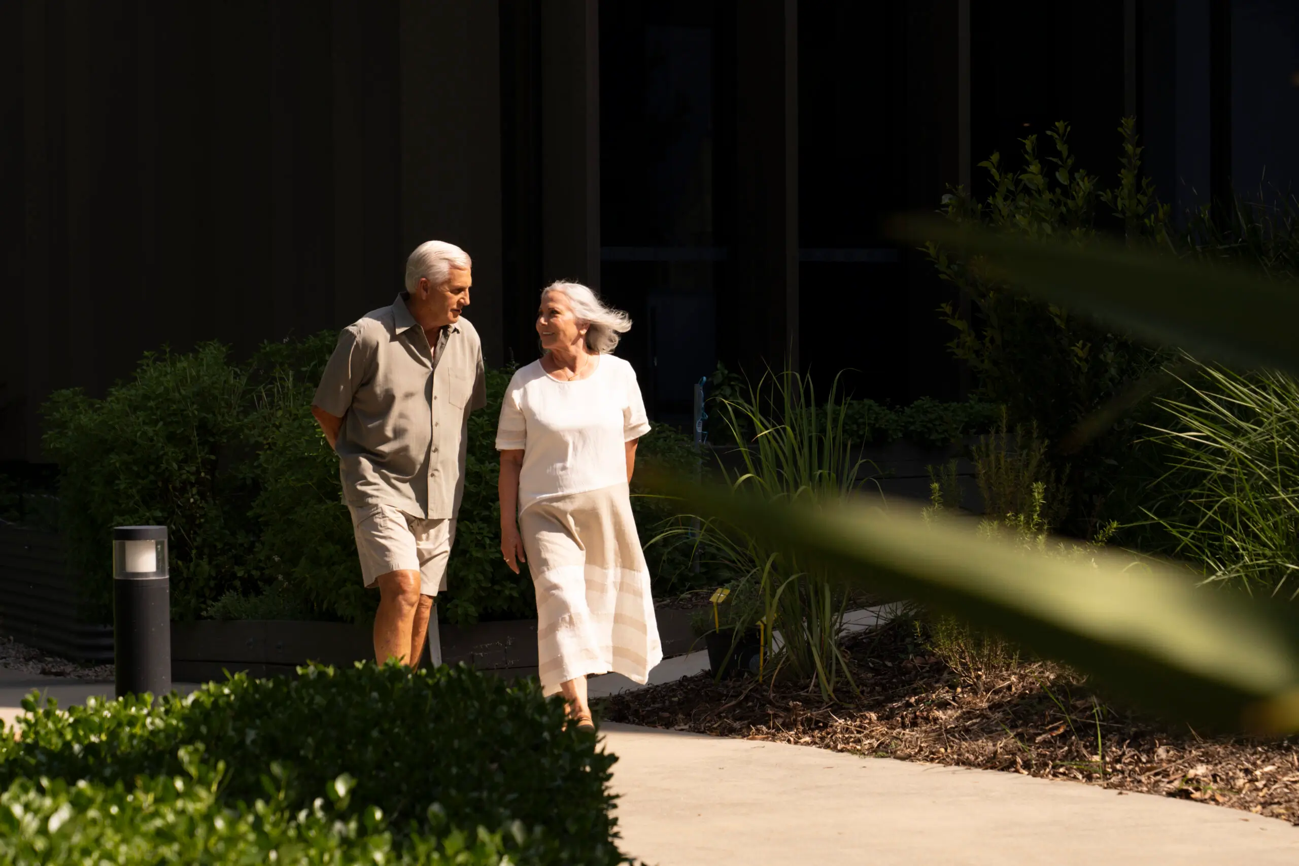 Two older adults, a man and a woman, walk together outdoors on a sunny day at The Evelyn Mt Ommaney, smiling at each other amid lush greenery and plants, both dressed in light, casual clothing.