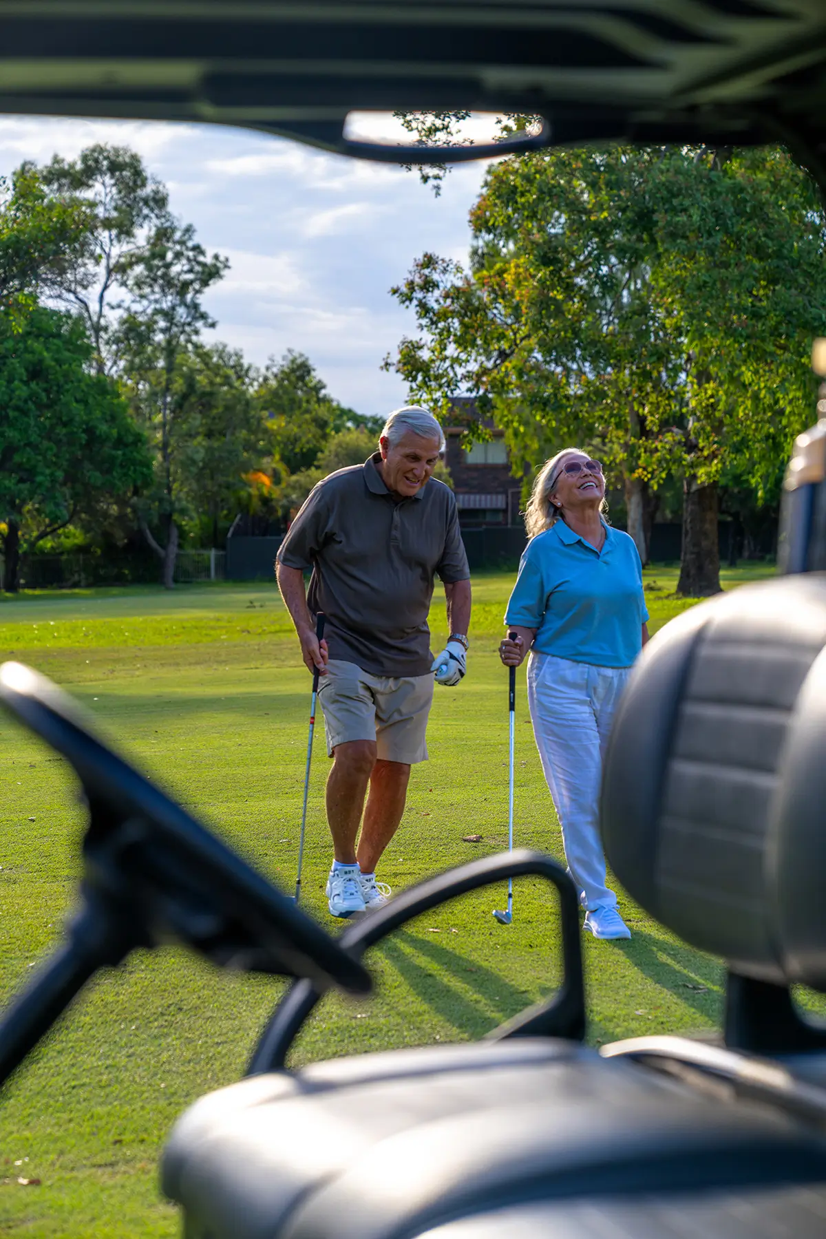 An older man and woman, both holding golf clubs, smile and walk together on the sunny course at The Evelyn Mt Ommaney, viewed from behind a golf cart with green trees in the background.
