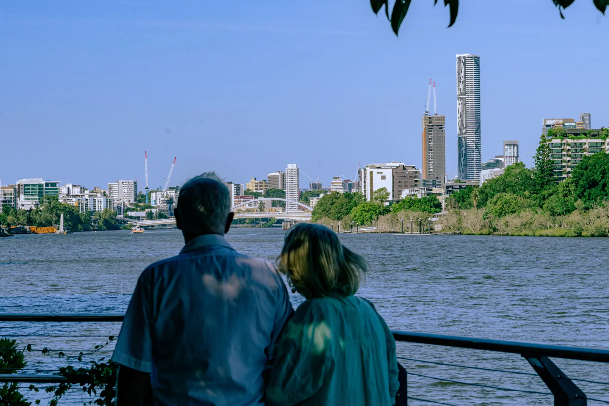 Two people stand by a river near the Milton retirement community, looking at a city skyline with tall buildings, trees, and a clear blue sky in the background. The scene appears calm and peaceful.