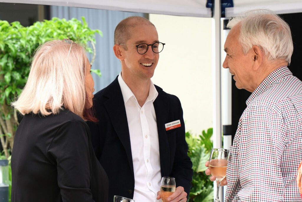 Three people stand together outdoors, holding drinks and smiling while conversing. One man wears glasses and a suit with a name tag, while the other two, a woman and an older man, are casually dressed. Greenery is visible in the background.