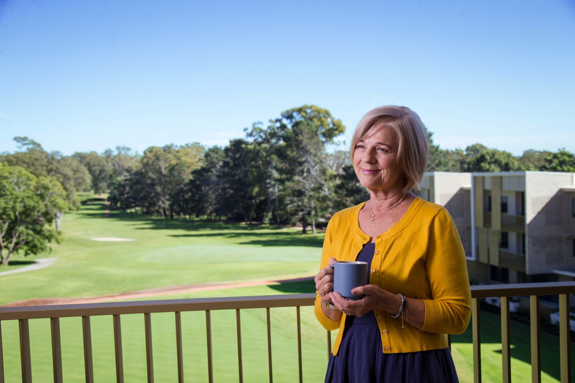 A woman with short blonde hair, wearing a yellow cardigan, stands on a balcony holding a mug. Behind her is a green golf course, trees, and a building, all under a clear blue sky.