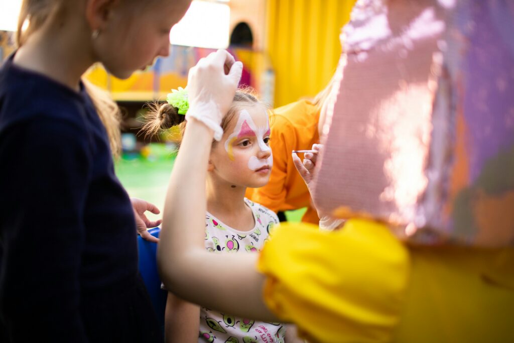 A young girl sits still as her face is painted with colorful designs at a Somerset Indooroopilly children's event. Other children and adults, one in a shiny costume, stand around her in a brightly lit, festive room.