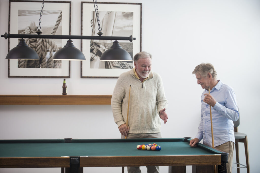 two older men, wearing a beige sweater and a light blue dress shirt talking to each other standing next to a pool table