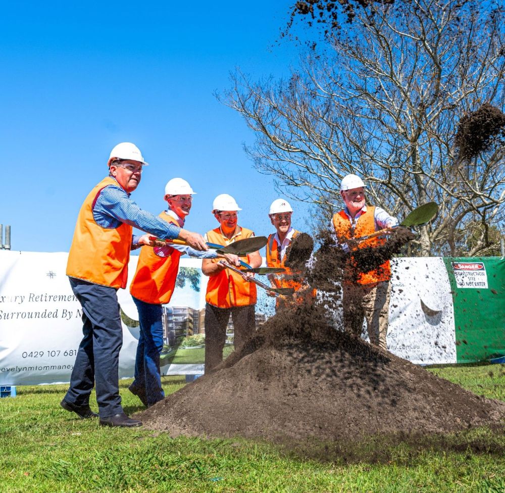 Five people in orange safety vests and white hard hats are smiling and tossing dirt with shovels at a groundbreaking ceremony for new pet friendly retirement villages, outdoors under a clear blue sky. A banner and trees are visible in the background.