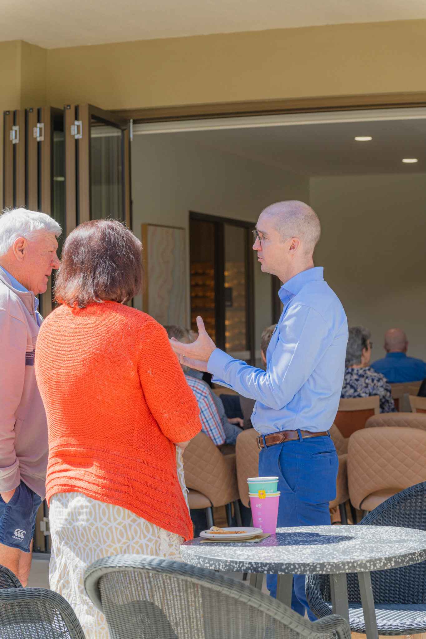 A man in a blue shirt speaks to two older adults, one in an orange sweater, outside a building with people seated inside. A purple coffee cup sits on a metal outdoor table in the foreground.