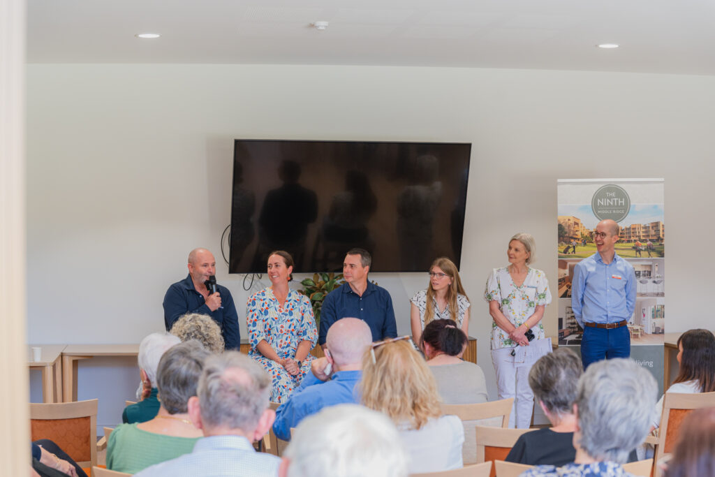 A panel of six people sits and stands at the front of a room, speaking to an audience. A man holds a microphone, and a TV and promotional banner are visible behind them. The audience is seated and facing the panel.