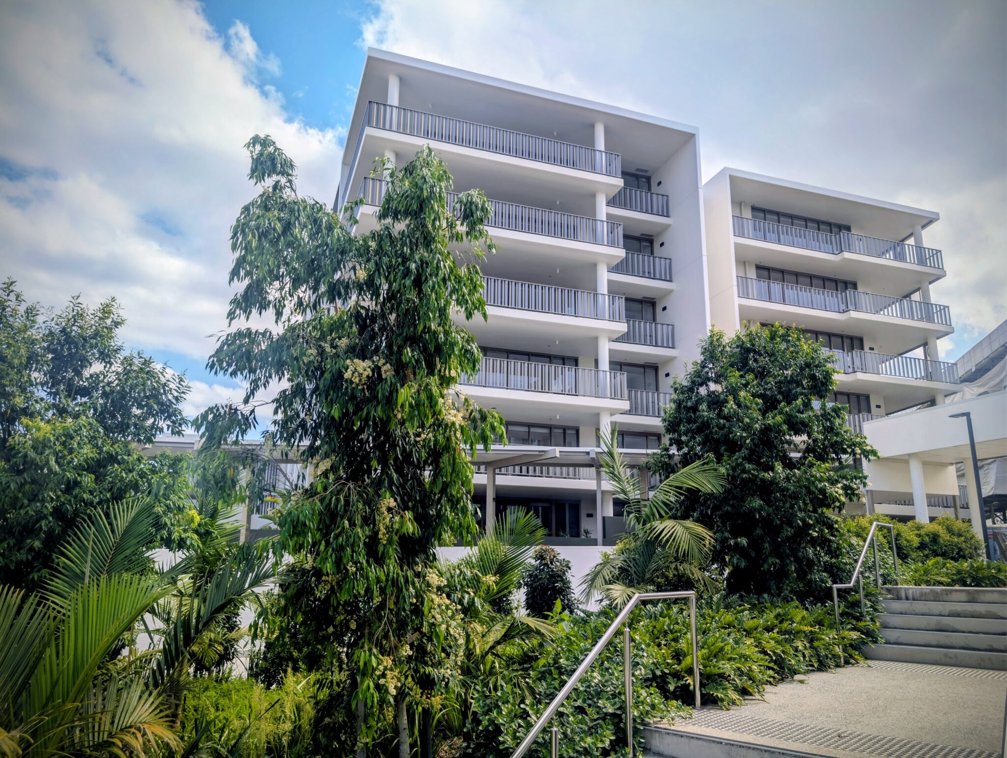 A modern white apartment building with multiple balconies is surrounded by lush green trees and plants, with a stairway and metal railing in the foreground under a partly cloudy sky.