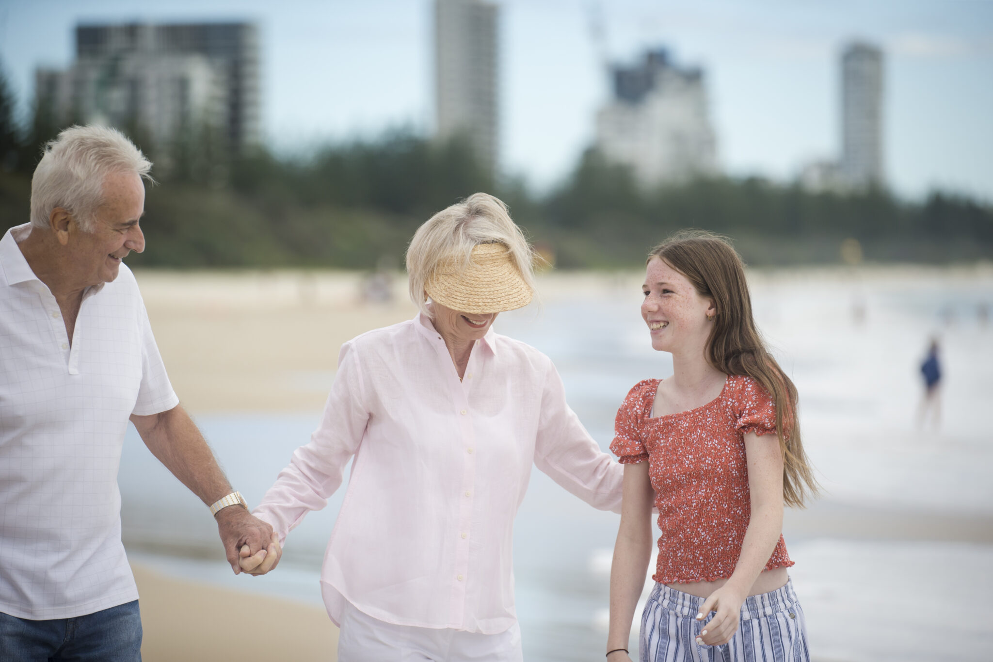 An older couple and a young girl walk along a sandy beach, smiling and holding hands, enjoying the joys of retirement living with tall buildings and trees in the background.