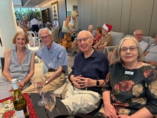 Four older adults, two men and two women, are sitting closely together at a table with empty wine glasses and a wine bottle. People are gathered in the background, one wearing a Santa hat, in a bright modern indoor space.