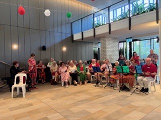 A group of elderly people sit in a semi-circle indoors, some holding musical instruments and sheet music, while others watch. Festive decorations hang from the ceiling. A woman stands and appears to be leading the group.