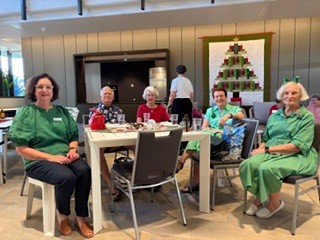 Five older adults sit around a table in a bright room, smiling at the camera. Some wear green outfits. A large quilted Christmas tree decorates the back wall. Drinks and small items are on the table.