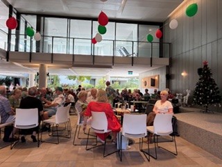A group of people sit at tables in a bright, modern room decorated with red, green, and white balloons. A Christmas tree stands in the corner, and people are chatting and enjoying refreshments.