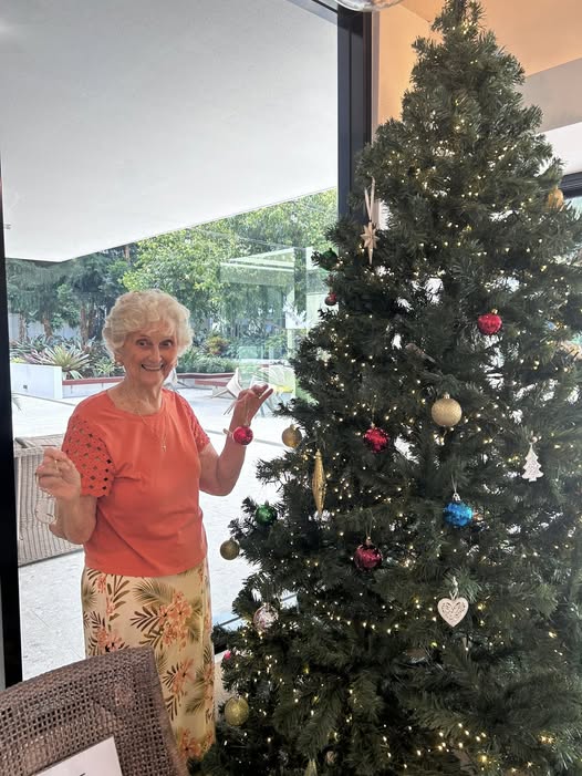 An elderly woman with white hair, wearing an orange top and floral skirt, smiles while helping decorate a lit Christmas tree with ornaments indoors next to a large window.