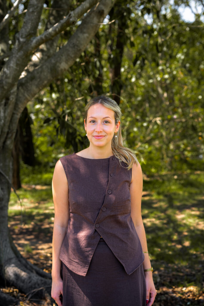 A woman stands outdoors in front of a large tree on a golf course, wearing a brown sleeveless top with buttons and a matching skirt. Sunlight filters through the leaves, capturing the serene ambiance of golf course living.