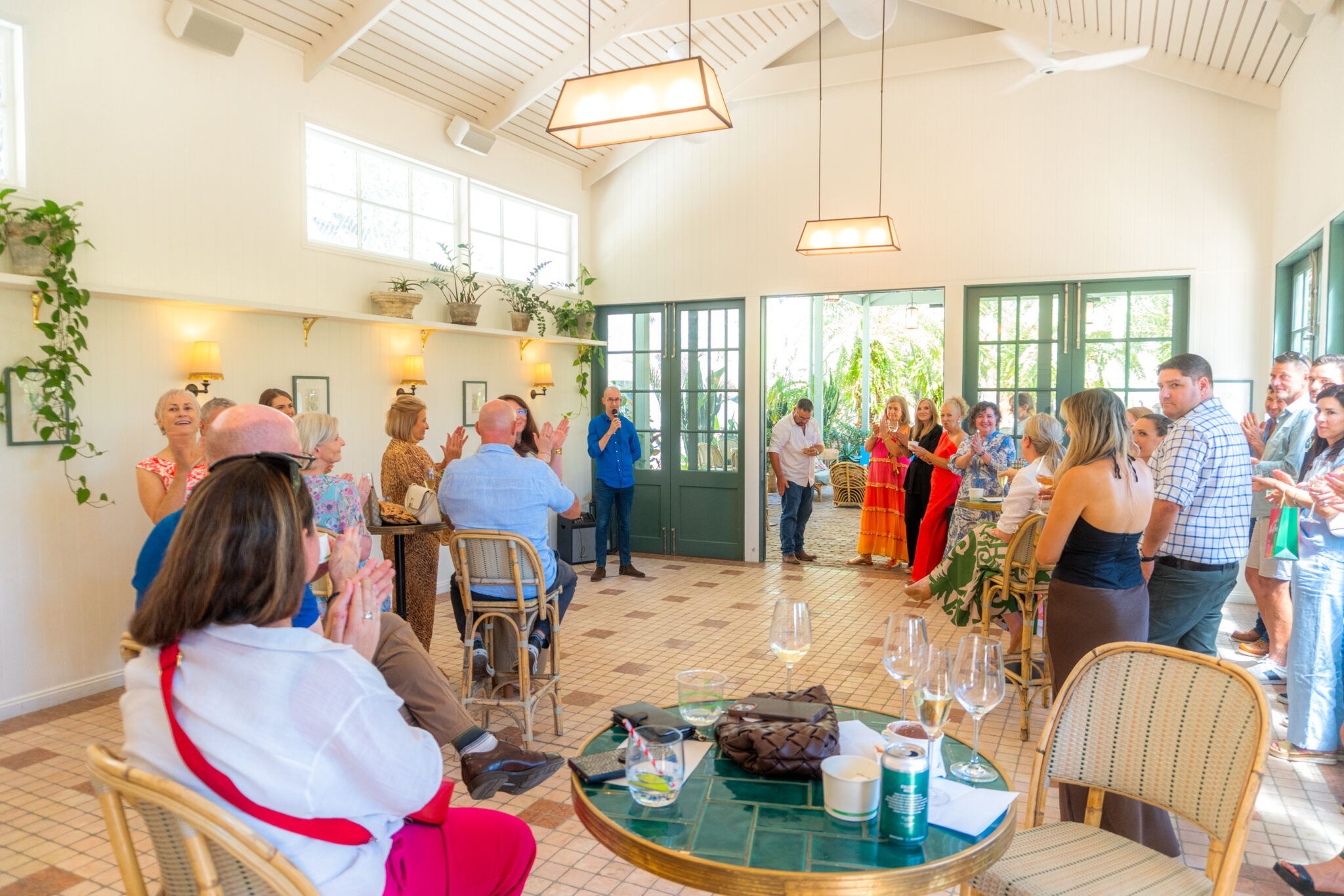 A group of people stand and sit in a bright room with large windows, clapping and watching a speaker in blue at the front. Tables have drinks and bags, and plants hang on the walls.