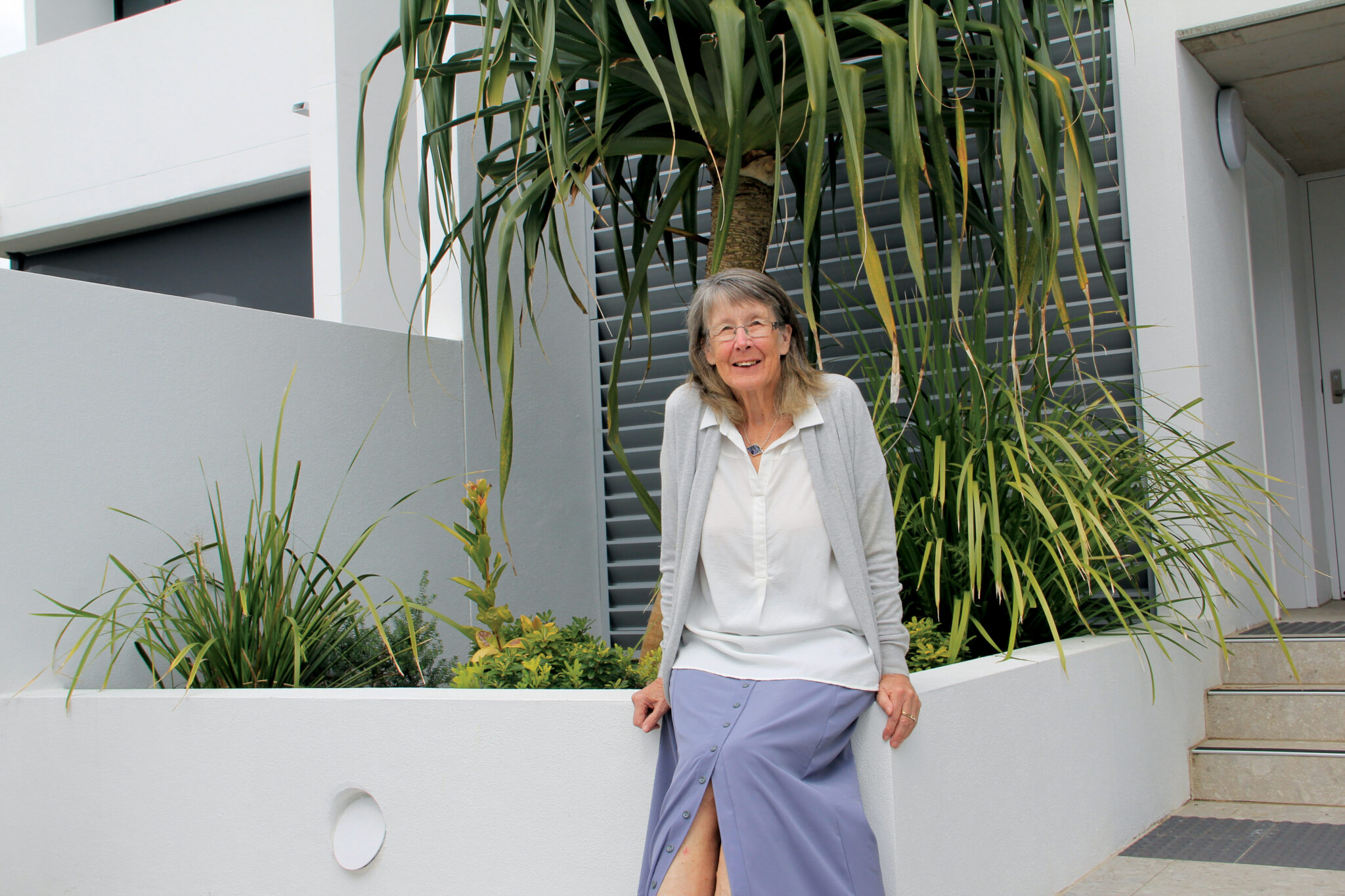 An older woman with glasses, long hair, and a gentle smile sits on a white garden wall in front of modern architecture and green plants—an inviting scene that captures the ease and comfort of what retirement living can offer.