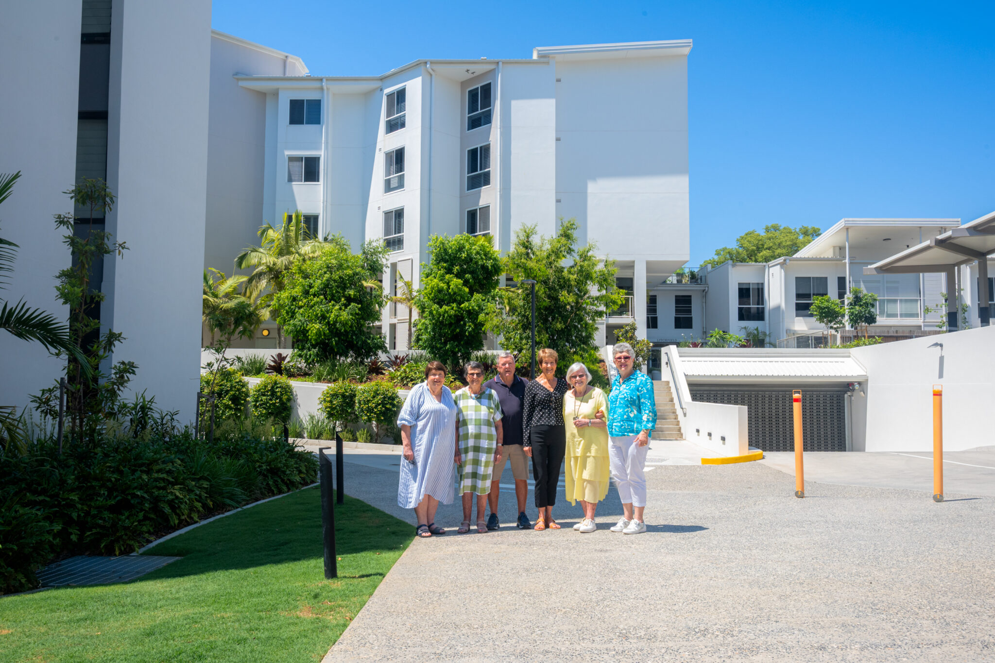 Five older adults stand together smiling on a sunny day in front of modern white apartment buildings with green landscaping and clear blue sky.