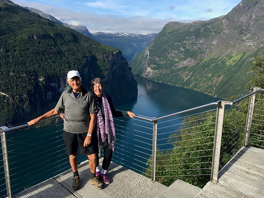 An older couple stands smiling on a viewing platform, embodying what is retirement living as they take in a deep river valley, surrounded by steep green mountains and blue water beneath a partly cloudy sky.