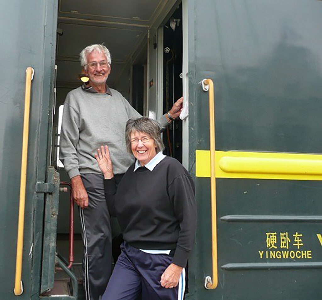 An older man and woman smile while standing on the steps of a green train carriage labeled "YINGWOCHE." Their joyful moment reflects what retirement living can look like—adventure, companionship, and new journeys together.