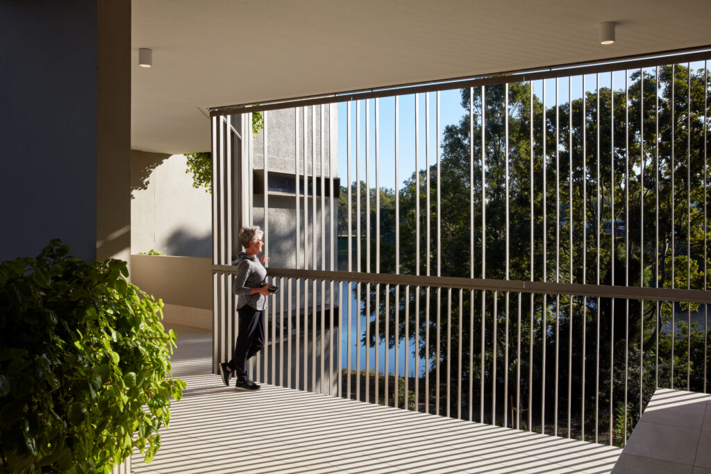 A person stands on a modern balcony with vertical metal railings in an award-winning Somerset residence, gazing at leafy trees as sunlight casts striped shadows on the floor.