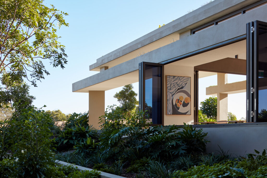 A modern, award-winning Somerset house with large open glass doors is surrounded by lush green plants and trees. A colorful abstract painting is visible inside on the wall, beneath a clear blue sky above the building.