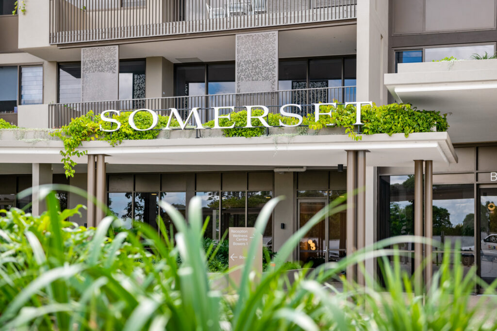 The entrance of a modern building with "SOMERSET" in large white letters above the doorway, surrounded by greenery and plants in the foreground.