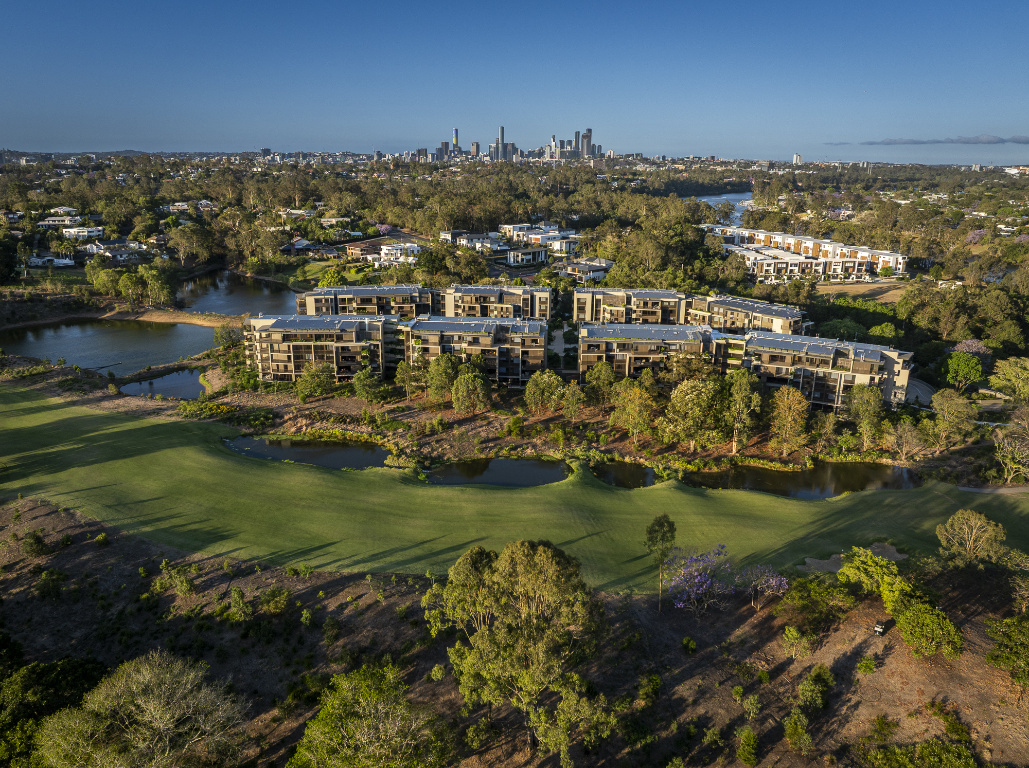 Aerial view of a modern apartment complex surrounded by trees and grassy areas, with a golf course in the foreground and a city skyline in the distance under a clear blue sky.