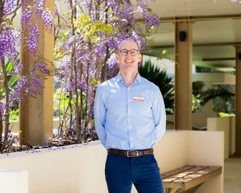 A person wearing glasses, a light blue shirt, and dark blue pants stands smiling in a sunlit outdoor area with benches and purple flowering vines overhead. Petals are scattered on the ground.