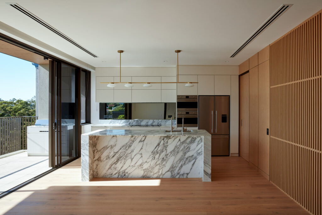 Modern kitchen with marble island and countertops, light wood floors, built-in appliances, and minimalist cabinetry. Large sliding glass doors open to a balcony with outdoor grill and scenic views.