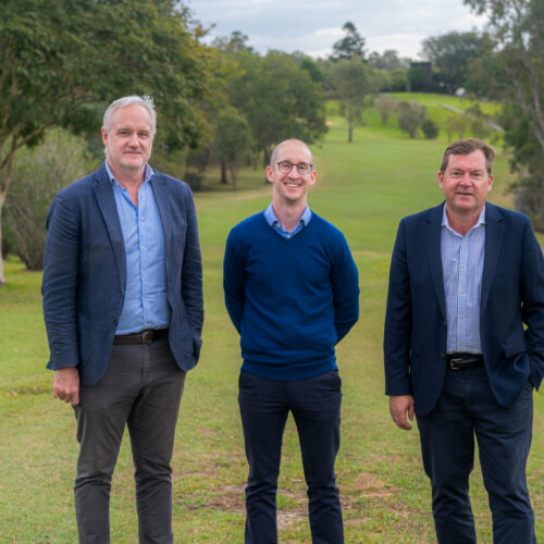 Three men stand side by side on a grassy field with trees and hills in the background. Smiling at the camera in business casual attire, they celebrate as Stonepeak announces investment in Aura Holdings.